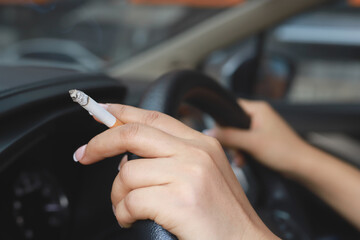 Hand holding burning cigarette while gripping steering wheel, with smoke filling car.