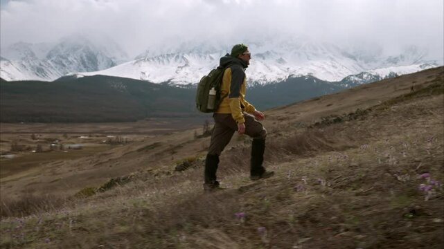 Male trekker with backpack climbing uphill against background of valley and snowy ridge in Altai mountains. Solo mountain hike on cloudy spring day, side view