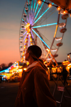 Young woman looking at illuminated Ferris wheel at night, vibrant fairground lights at dusk.