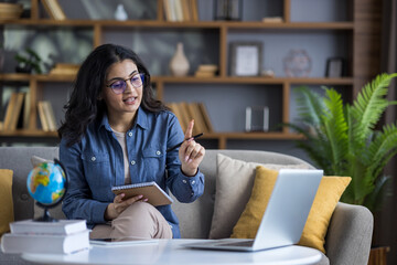 Young Indian female student sitting on the couch at home, holding a notebook and conducting distance learning via laptop
