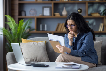 Serious young Indian woman sitting on sofa at home and counting bills, looking at receipts and...