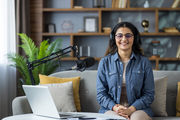 Portrait of a young Indian woman in a headset sitting on a sofa at home in front of a microphone and looking at the camera