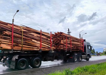 large truck is carrying significant load logs on highway surrounded by greenery, with gray clouds...