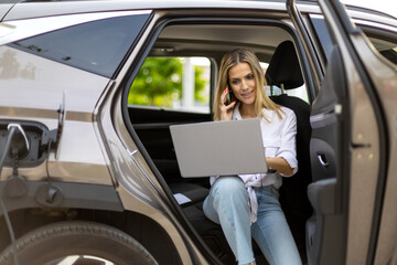 Businesswoman working on laptop and talking on phone while charging electric car