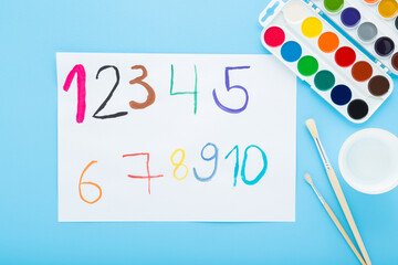 Colorful watercolor palette, paintbrushes and drawn different numbers on white paper on light blue table background. Pastel color. Closeup. Creativity learning for little kids. Top down view.