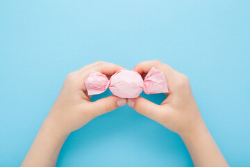 Little child girl hand fingers holding and showing candy wrapped in pink paper on light blue table background. Pastel color. Closeup. Point of view shot. Top down view.