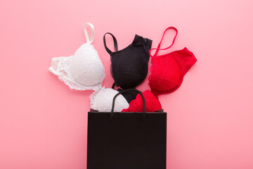 Shopping paper bag with new white, black and red lace bras on light pink table background. Pastel...