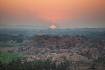 A serene sunrise or sunset over the ancient landscape of Hampi, India, with a large orange sun descending over rocky hills, lush palm trees, and distant fields