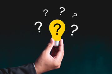 A hand holding a glowing yellow light bulb surrounded by multiple question marks, symbolizing the process of idea generation, problem-solving and seeking answers, innovation and strategic thinking.