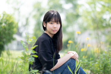 Young asian teenage girl sitting on the ground in a grassy area, surrounded by green plants and wildflowers, wearing a black top and jeans.