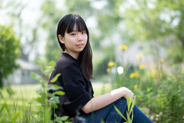 Young asian teenage girl sitting on the ground in a grassy area, surrounded by green plants and wildflowers, wearing a black top and jeans.
