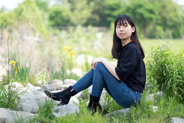 Young asian teenage girl sitting on the ground in a grassy area, surrounded by green plants and wildflowers, wearing a black top and jeans.