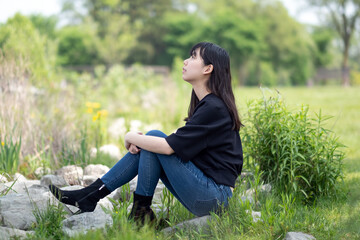 Young asian teenage girl sitting on the ground in a grassy area, surrounded by green plants and wildflowers, wearing a black top and jeans.