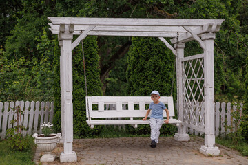 Little boy relaxing alone on a white wooden garden swing with rustic frame, enjoying calm childhood moments and simple outdoor leisure in a peaceful backyard.