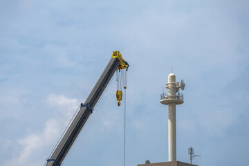 Construction crane positioned next to tall communication tower extends diagonally across light blue sky backdrop dotted with scattered fluffy clouds.