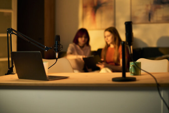 Two Caucasian teenage girls sitting together discussing content while preparing podcast episode in modern studio with microphones and laptop on table in foreground