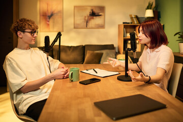 Two Caucasian teenagers recording podcast at table, boy wearing glasses listening while girl gesturing and speaking into microphone, smartphone and laptop on wooden surface