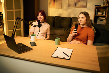 Two Caucasian teenage girls recording podcast sitting at table with microphones and laptop, both...