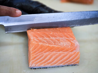 Fresh salmon fillet being sliced with a sharp knife by a chef wearing a black glove. High-quality seafood preparation in a professional kitchen setting.