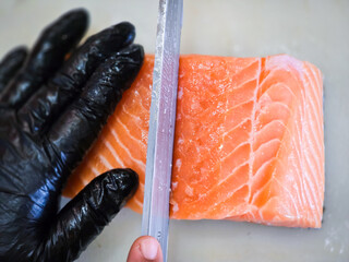 A chef slicing a fresh salmon fillet with a sharp knife. The chef wears black gloves for hygiene, preparing the fish on a clean white cutting surface.