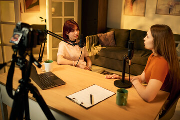 Two Caucasian teenage girls recording podcast conversation at table with microphones, laptop,...