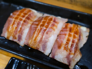Grilled bacon-wrapped sushi served on a black tray. Close-up image highlighting the juicy texture, glossy sauce, and savory Japanese fusion style presentation.