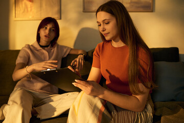 Two teenage girls sitting on sofa recording podcast, Caucasian girl holding clipboard and writing...