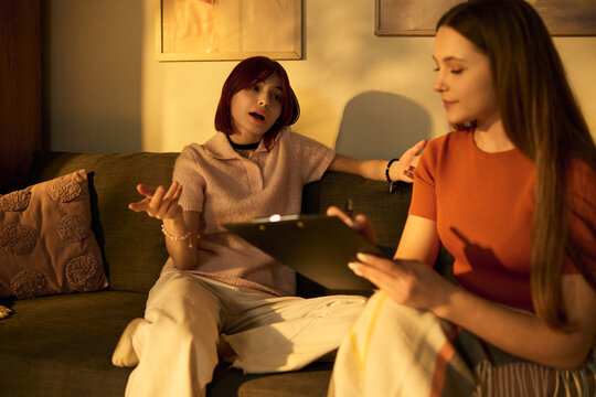 Two teenage girls sitting on sofa recording podcast episode, Caucasian girl holding clipboard listening attentively while Asian girl gesturing and speaking into microphone out of frame