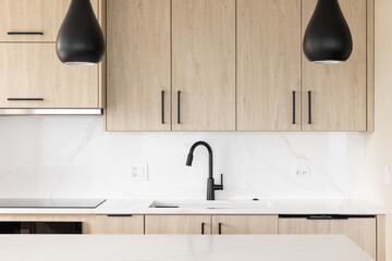 A kitchen detail with a black faucet on a white marble countertop, modern wood cabinets, and black...