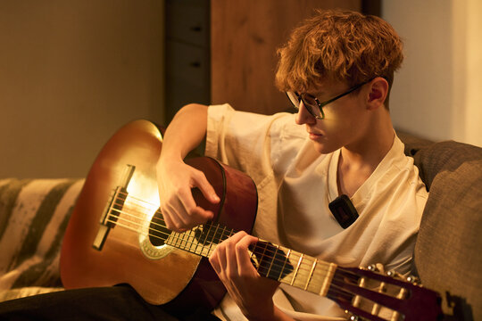 Caucasian teenage boy sitting on sofa playing acoustic guitar wearing glasses and microphone headset recording podcast content focused on music performance in home setting
