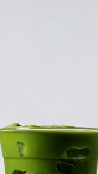 Close-up of matcha green tea being poured over ice in a transparent glass with rich texture and creamy swirl effect on white surface