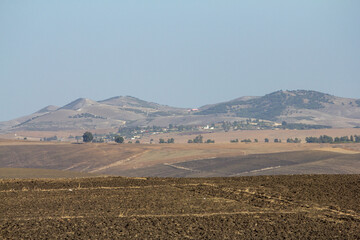 An expansive agricultural landscape features foreground fields of rich, dark brown tilled earth giving way to lighter, golden cultivated land, with a distant village nestled against rolling green hill