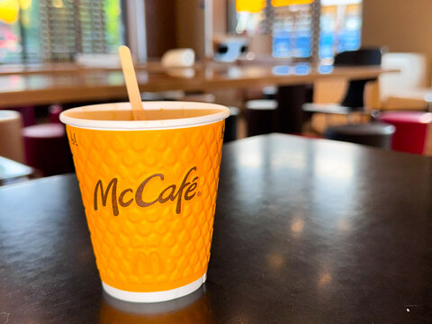 Vibrant orange disposable coffee cup with wooden stirrer placed on table in comfortable caf&eacute; atmosphere, showcasing warm and inviting ambiance for coffee lovers.