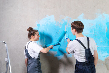 House decoration DIY renovation concept. Happy couple in empty room of new home painting wall together. Husband and wife holding paint roller painting wall with blue color paint in new house