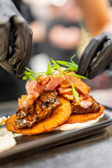 Close-up of a chef garnishing crispy potato pancakes with herbs, served with glazed meat and bacon. Professional food styling and elegant plating in restaurant kitchen.