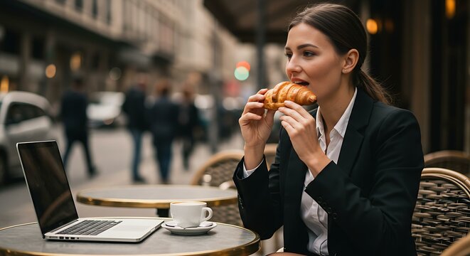 Businesswoman seated at an outdoor café table, taking a bite of a croissant - Powered by Adobe
