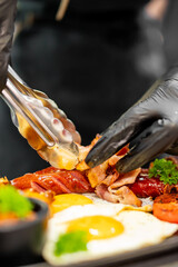 Close-up of chef's gloved hands arranging a hearty breakfast plate with fried eggs, bacon, sausage,...