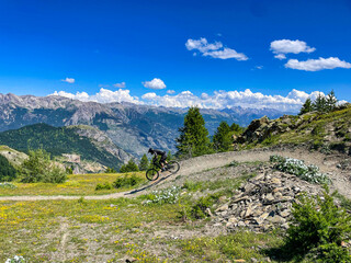 Mountain biker descending a winding dirt trail on a rugged alpine slope in the Alps, with majestic...