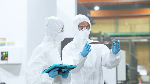Male and female workers in protective clothing holding tablets and inspecting silicon wafers in a semiconductor factory