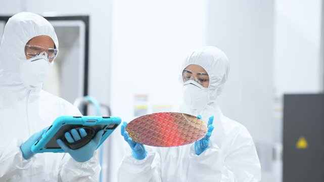Male and female workers in protective clothing holding tablets and inspecting silicon wafers in a semiconductor factory