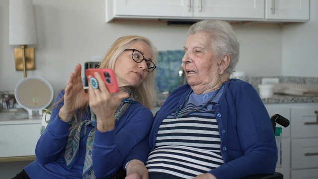 A daughter helps her elderly mother connect with family through a smartphone, capturing precious memories and fostering love and support in their cozy home. - Powered by Adobe