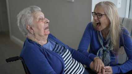 An elderly woman in her 90s smiles joyfully while seated in a wheelchair, sharing a warm moment with her daughter in a cozy indoor setting.