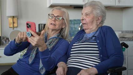 A woman in her 90s sits in a wheelchair while her daughter shows her something on a smartphone,...