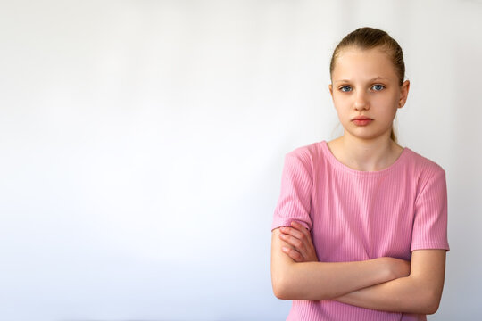 Upset Girl Standing with Arms Crossed Indoors. Serious young girl in pink shirt looking displeased or upset, standing with arms crossed against a plain white background. Space for text.