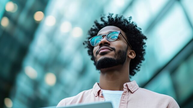 A thoughtful young man gazes upwards against a modern glass background, wearing glasses and a relaxed shirt, embodying contemplation and aspiration in his expression.
