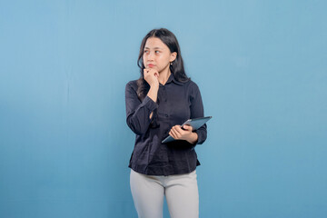 A woman is standing in front of a blue wall, looking at a tablet in her hand. She is deep in...