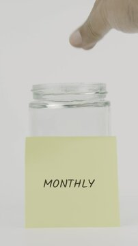 A yellow sticky note with the word monthly is attached to a glass jar as a hand inserts a coin. This vertical video reflects saving and budgeting on a consistent monthly basis