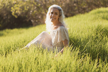 A cheerful woman sits in a sunlit meadow, surrounded by lush green grass. Her relaxed and joyful...