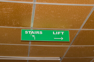green ceiling-mounted indicator board displaying directional arrows for stairs (left) and lift (right), commonly seen in hospitals, malls, office buildings, and public infrastructure