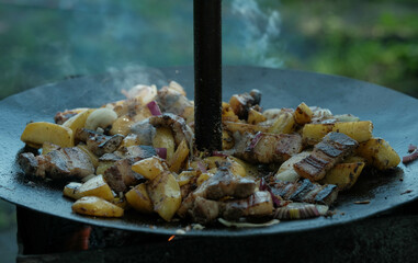Savory outdoor-cooked meal of fried potatoes, bacon, and onions, sizzling on a black metal disc.
